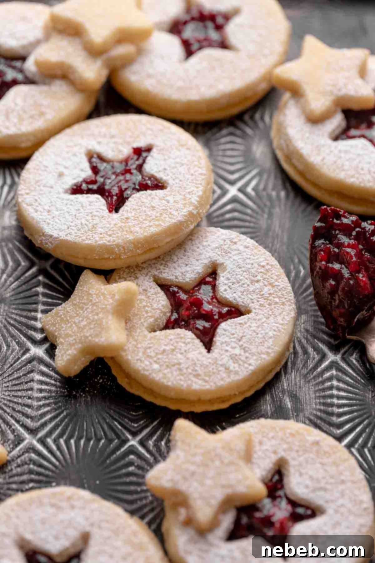 Beautifully arranged cranberry raspberry Linzer cookies, dusted with powdered sugar, on a dark tray. The top cookie reveals a festive cutout, showing the rich red jam filling.