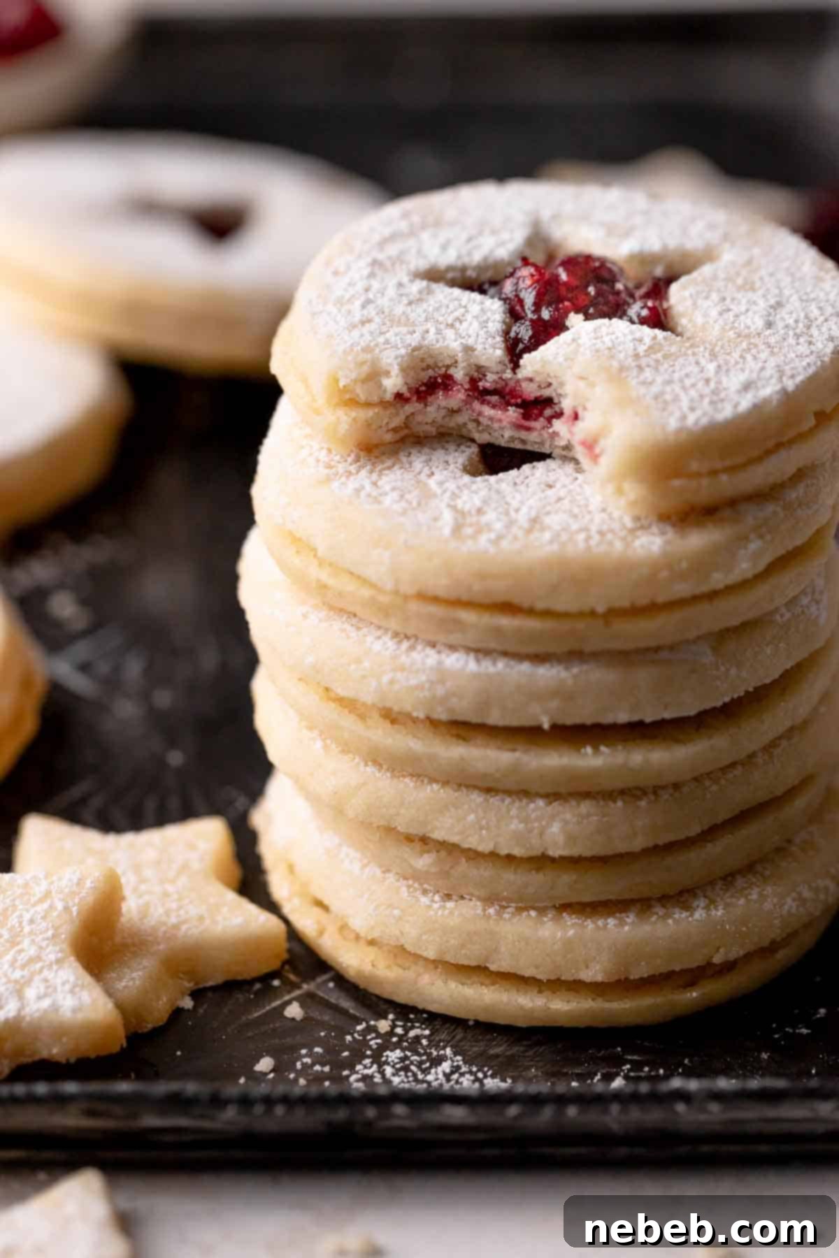 A delightful stack of raspberry Linzer cookies, showcasing their jam filling and powdered sugar dusting, resting on a sleek black baking tray.
