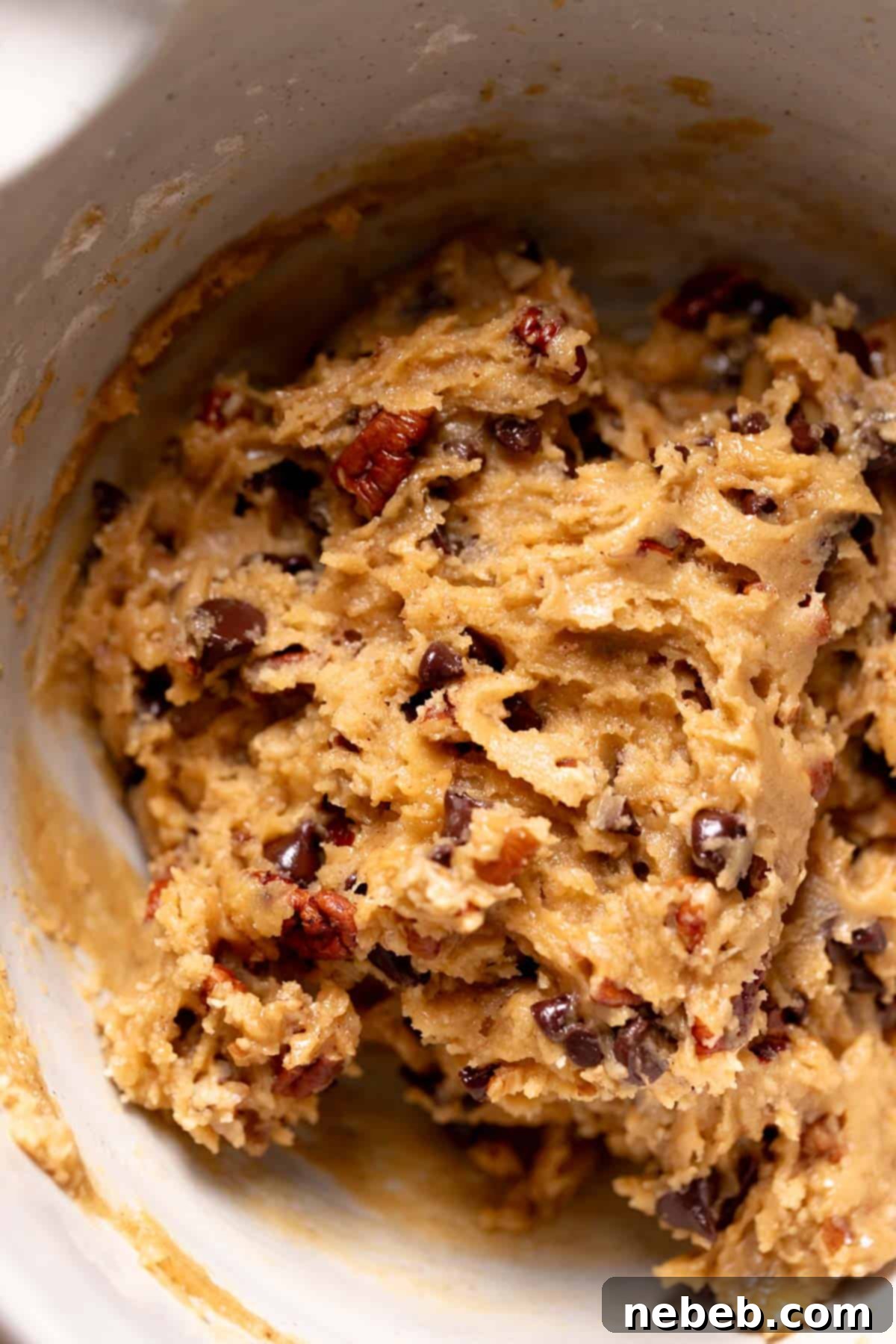 Rich chocolate chip pecan cookie dough in a large mixing bowl, showing chopped pecans and two types of chocolate chips distributed throughout.