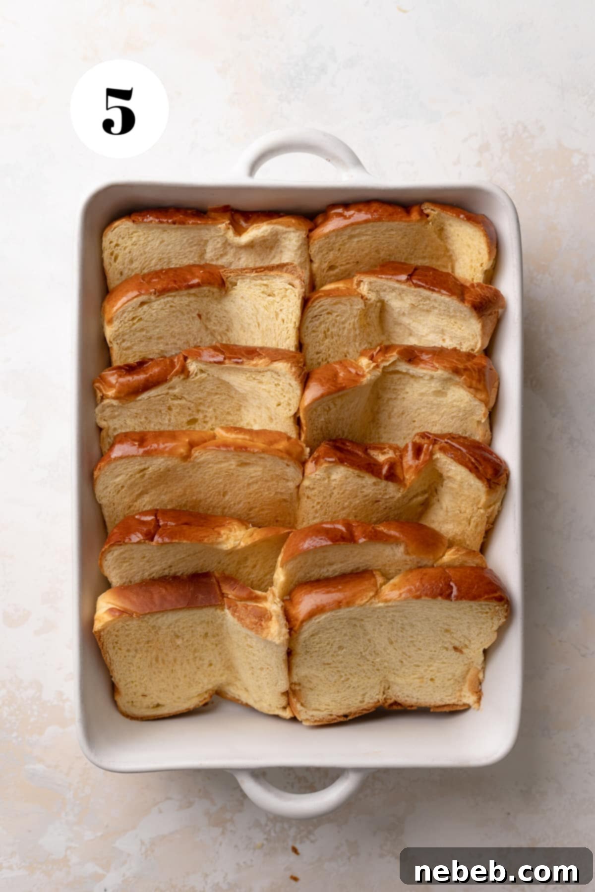Toasted brioche bread slices arranged in a domino pattern in a white baking dish, ready for custard.