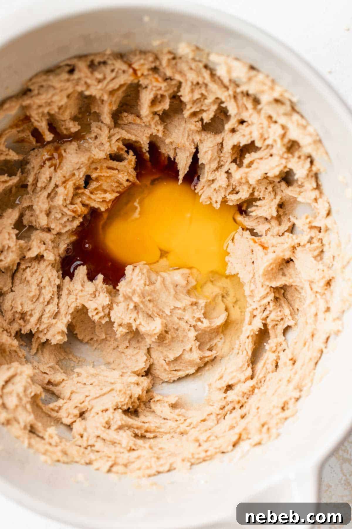 A mixing bowl with creamed butter and sugar with an egg cracked into the center of the bowl, ready to be mixed in.