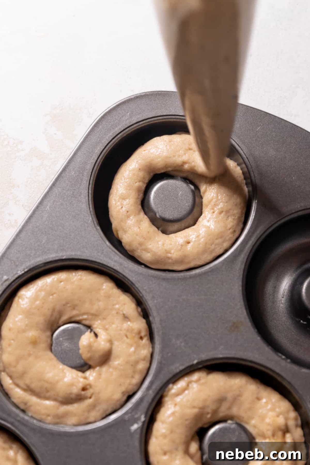 Piping the banana donut batter into a greased donut pan, ensuring even filling.