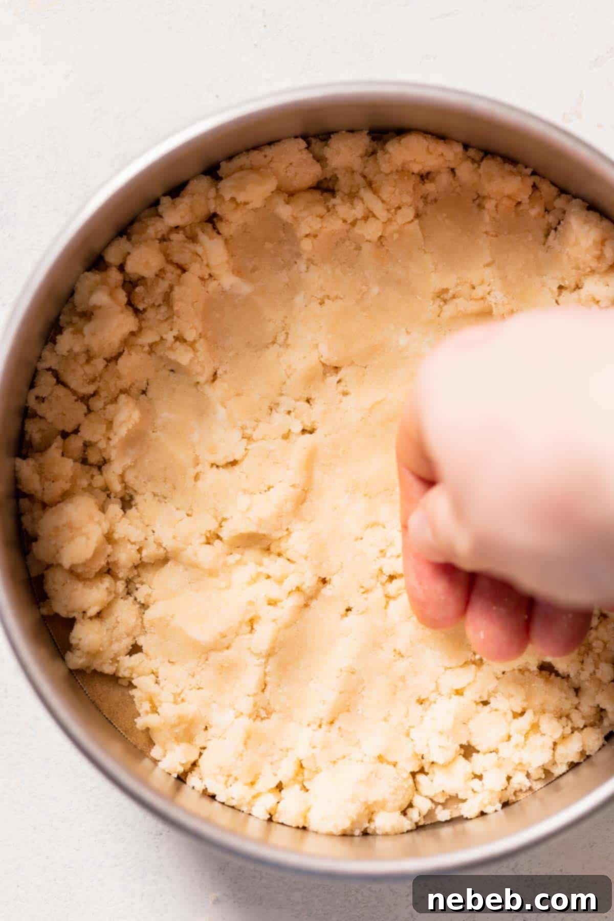 A hand firmly pressing the shortbread crumb mixture into the bottom of a round cake pan, forming an even crust.