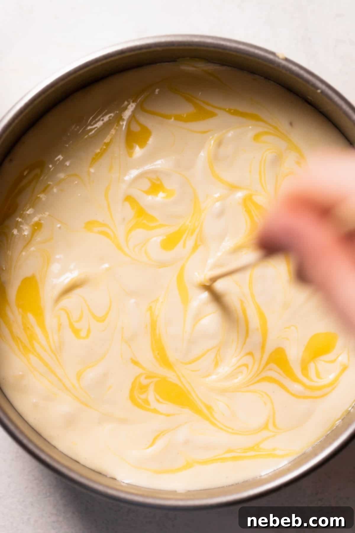 A toothpick gently swirling dollops of lemon curd into the cake batter within a round cake pan, creating a marbled effect.