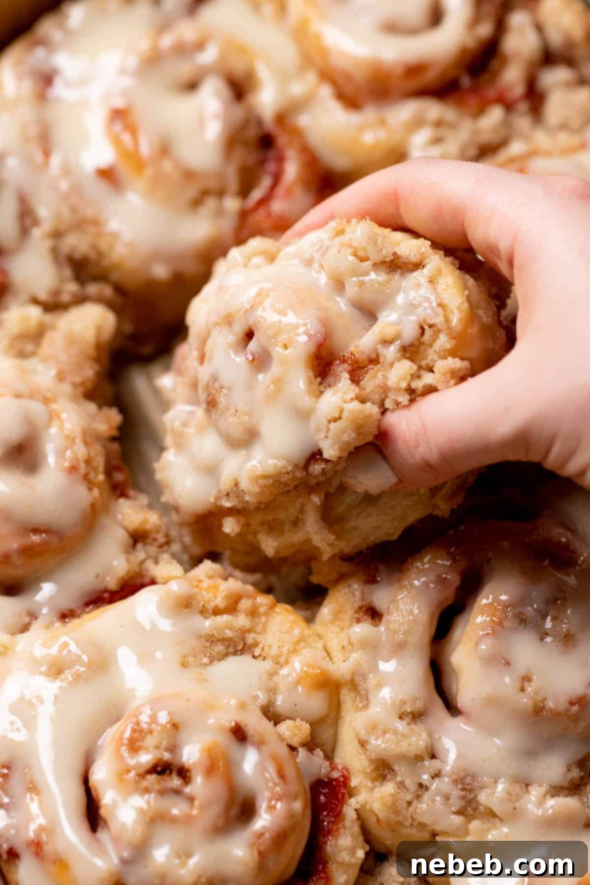A hand reaching into a baking pan to pull out a single peach cobbler cinnamon roll, still warm and glazed.