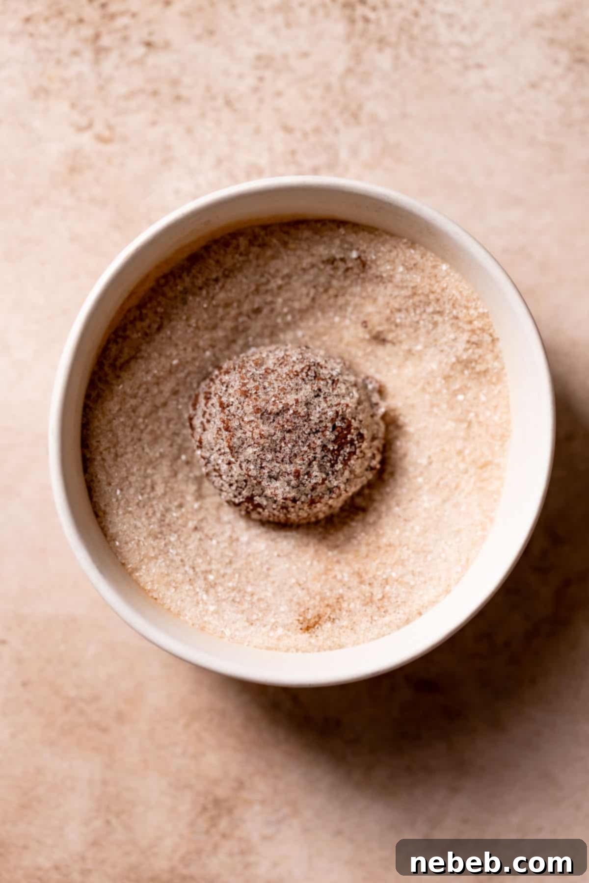 A chocolate cookie dough ball being rolled in a bowl of cinnamon sugar, ensuring a thorough coating.