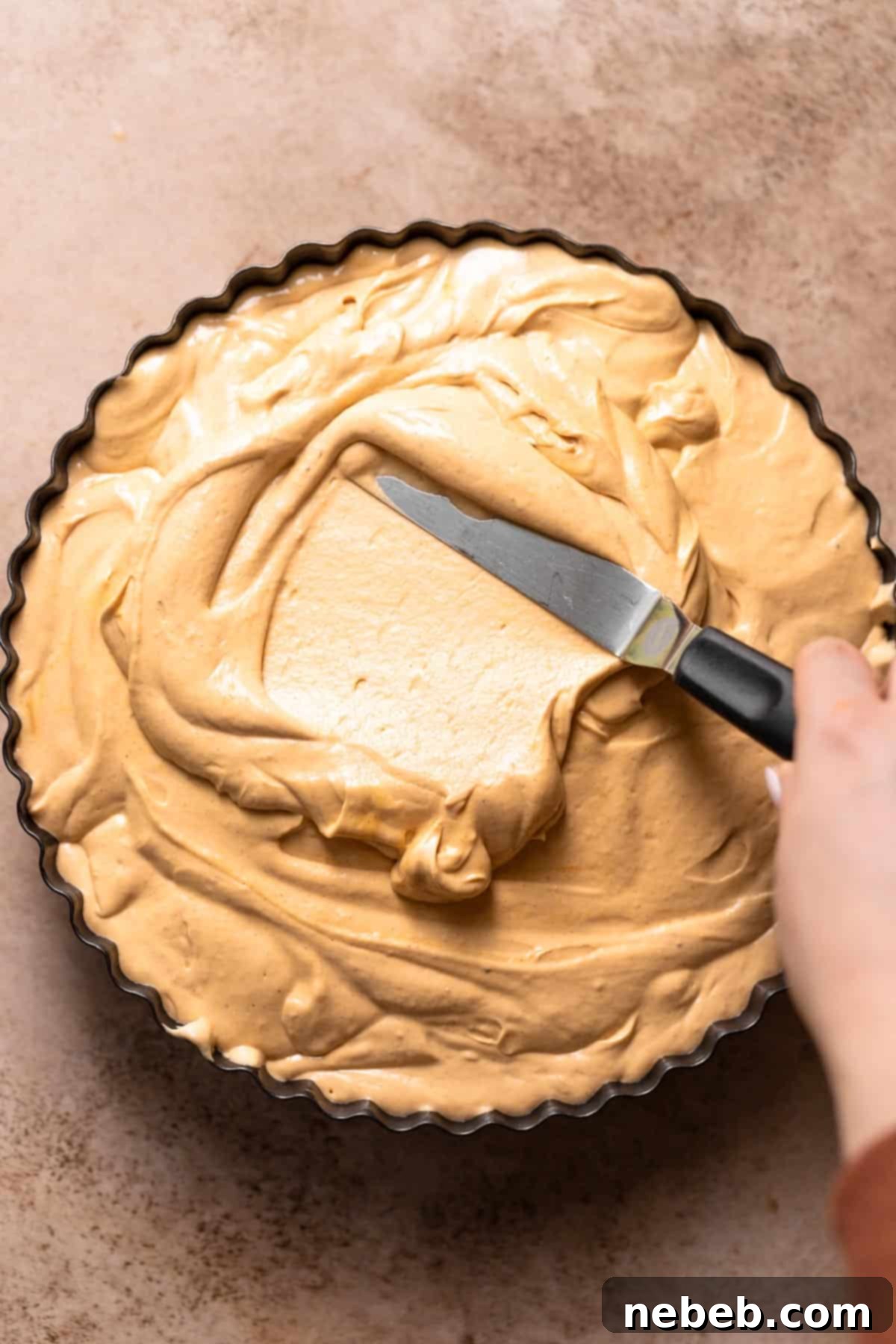 A hand using an offset spatula to carefully spread the creamy peanut butter filling into the prepared cake pan over the chocolate base.