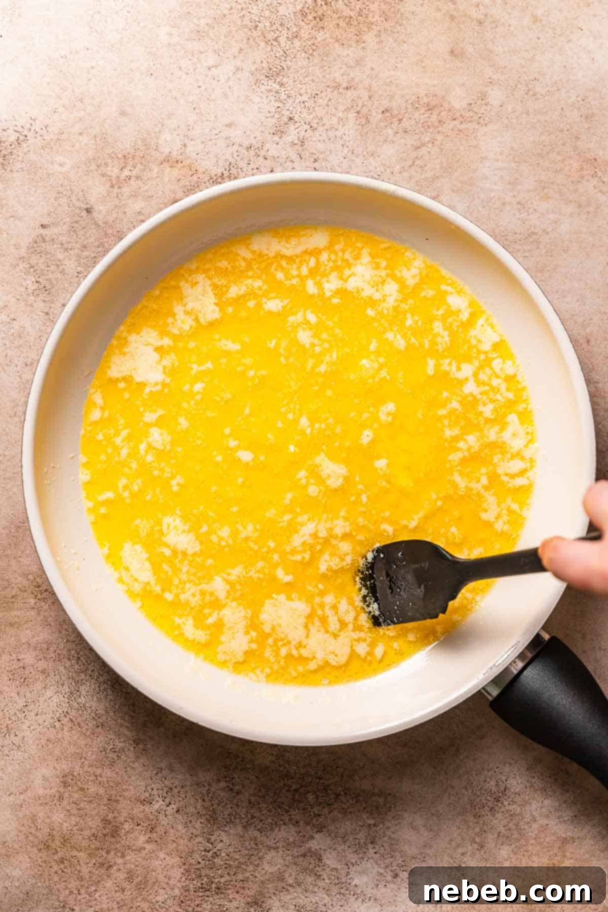 A white pan with bubbling butter being stirred with a black rubber spatula, actively browning.