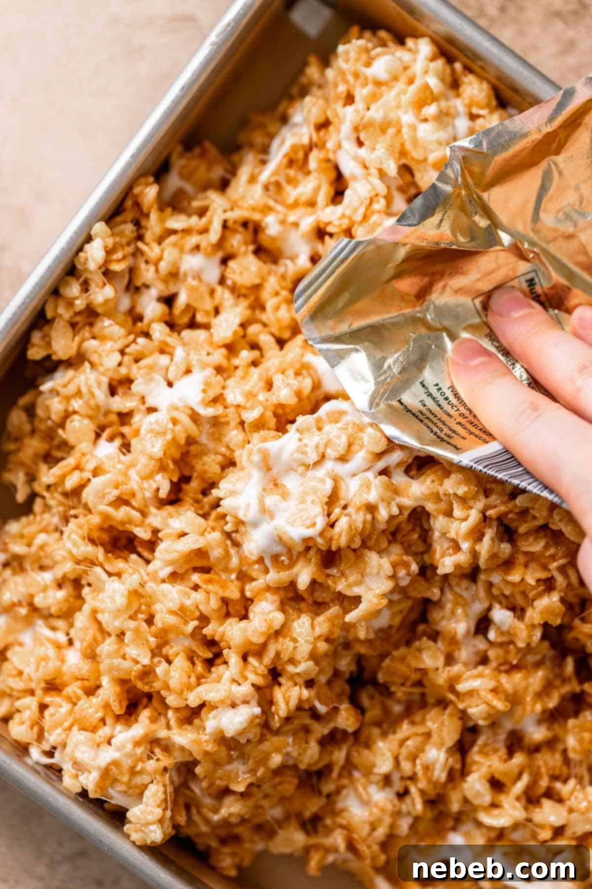 A hand using a butter wrapper to press the rice krispie mixture into the prepared pan.