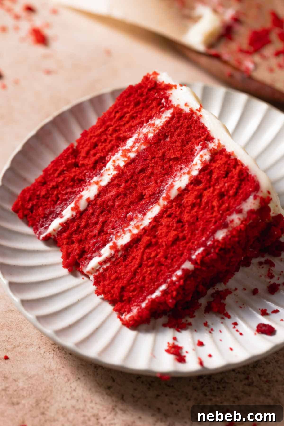 A beautifully presented slice of super moist red velvet cake on a pristine white plate, adorned with creamy white frosting and a sprinkle of red cake crumbs.