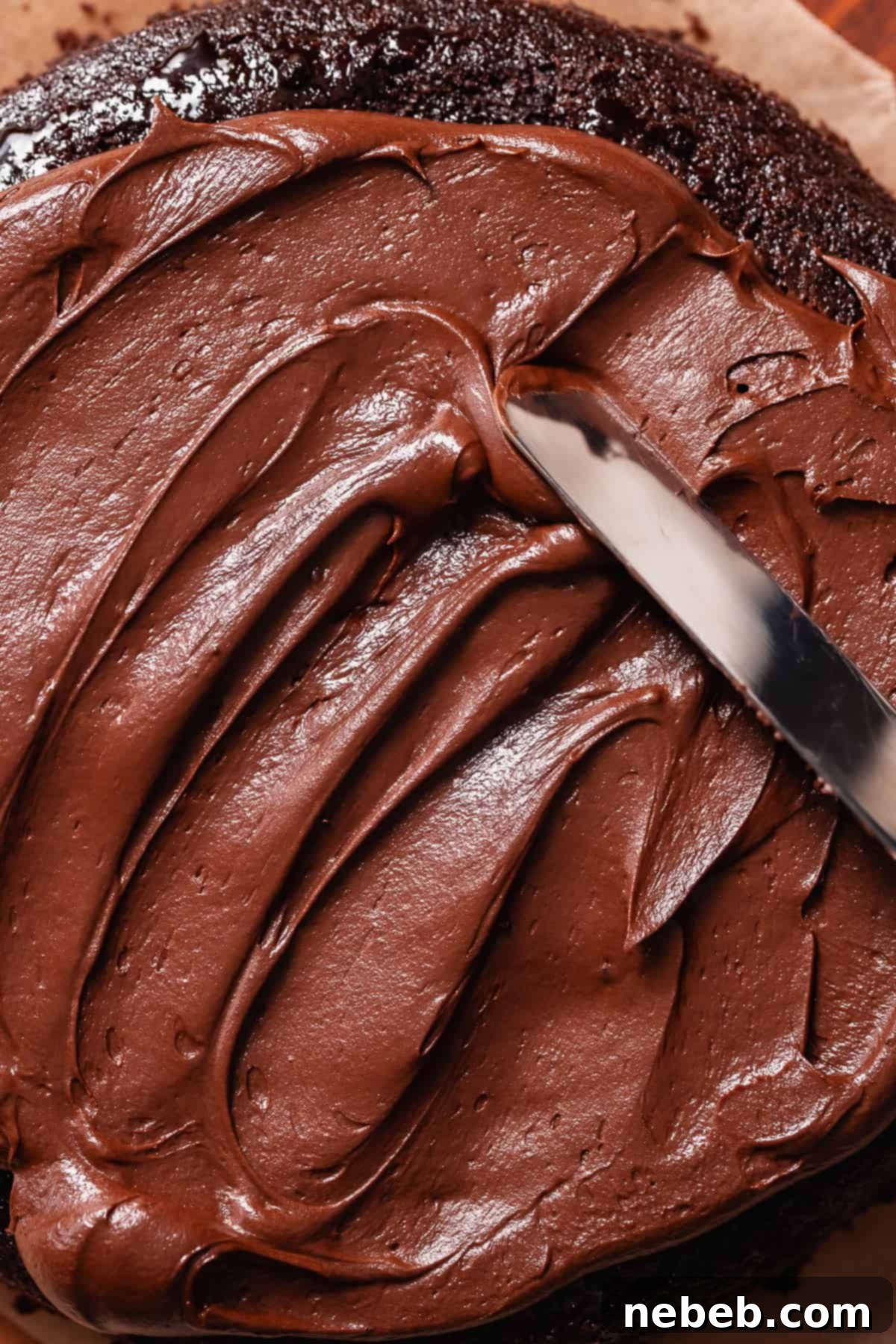 A spatula spread the chocolate buttercream frosting on a chocolate cake layer.