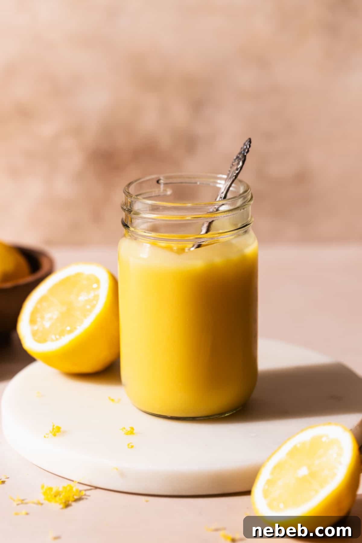 A glass mason jar of homemade lemon curd on a white marble stand with a spoon dipping into it, showcasing its creamy texture.