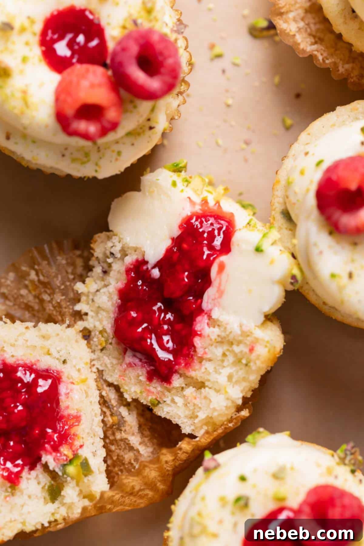 Two lemon cupcakes with raspberry filling cut in half, revealing the vibrant red jam inside, arranged on a baking tray alongside whole fresh raspberries and pistachios.