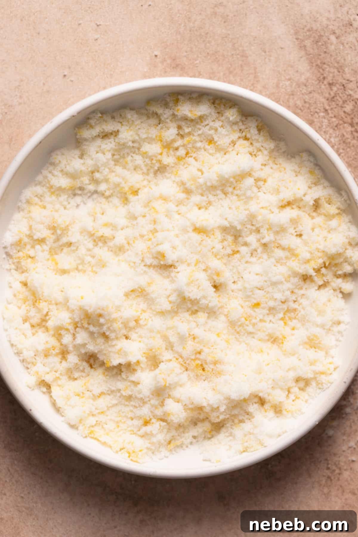 Close-up of lemon zest being rubbed into granulated sugar in a white mixing bowl, creating aromatic lemon sugar.