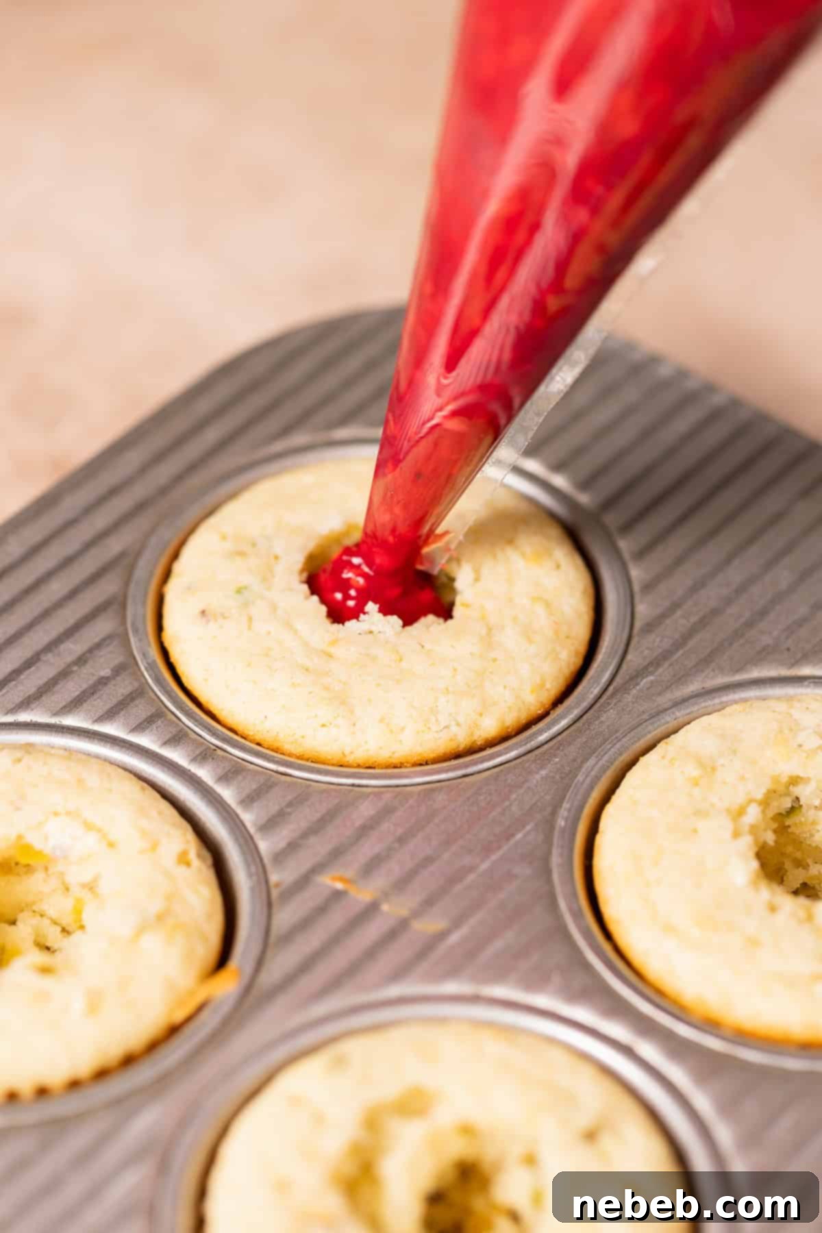A piping bag filling the hollowed-out center of a lemon cupcake with vibrant red raspberry jam.