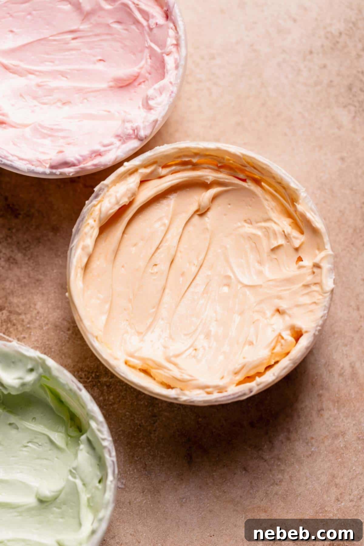 Three bowls with orange, green, and pink Swiss meringue buttercream, vibrantly colored and smooth.
