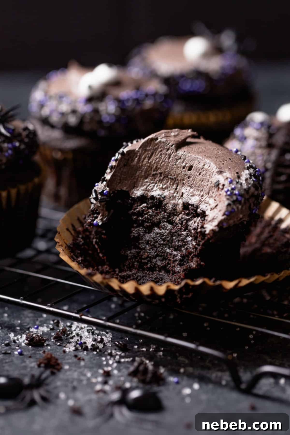 A dark black cupcake with a bite taken out of it, sitting on a black cooling rack. The frosting is a deep black color with festive sprinkles.