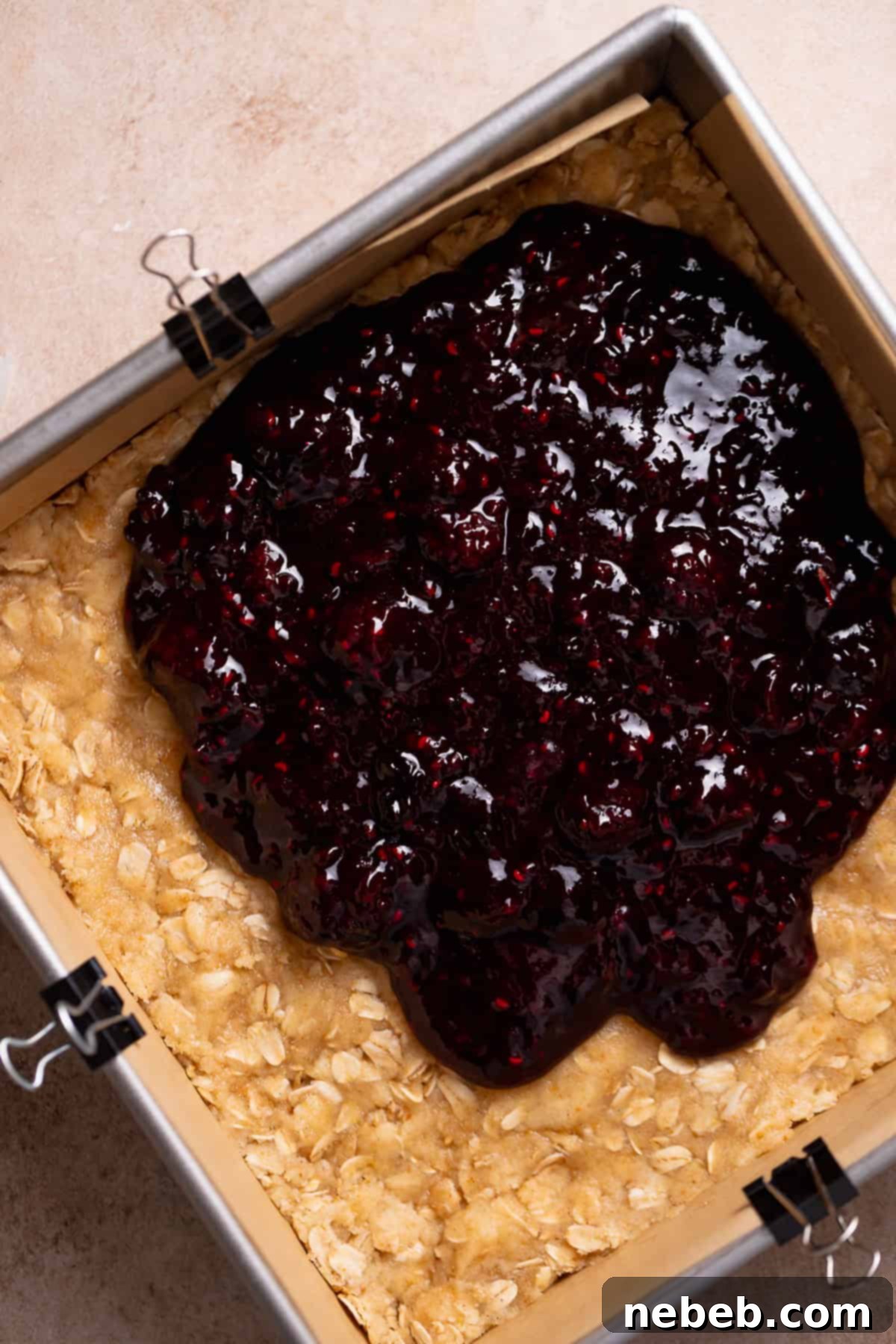 Mixed berry jam being spread over the oat crumble base in a baking pan.