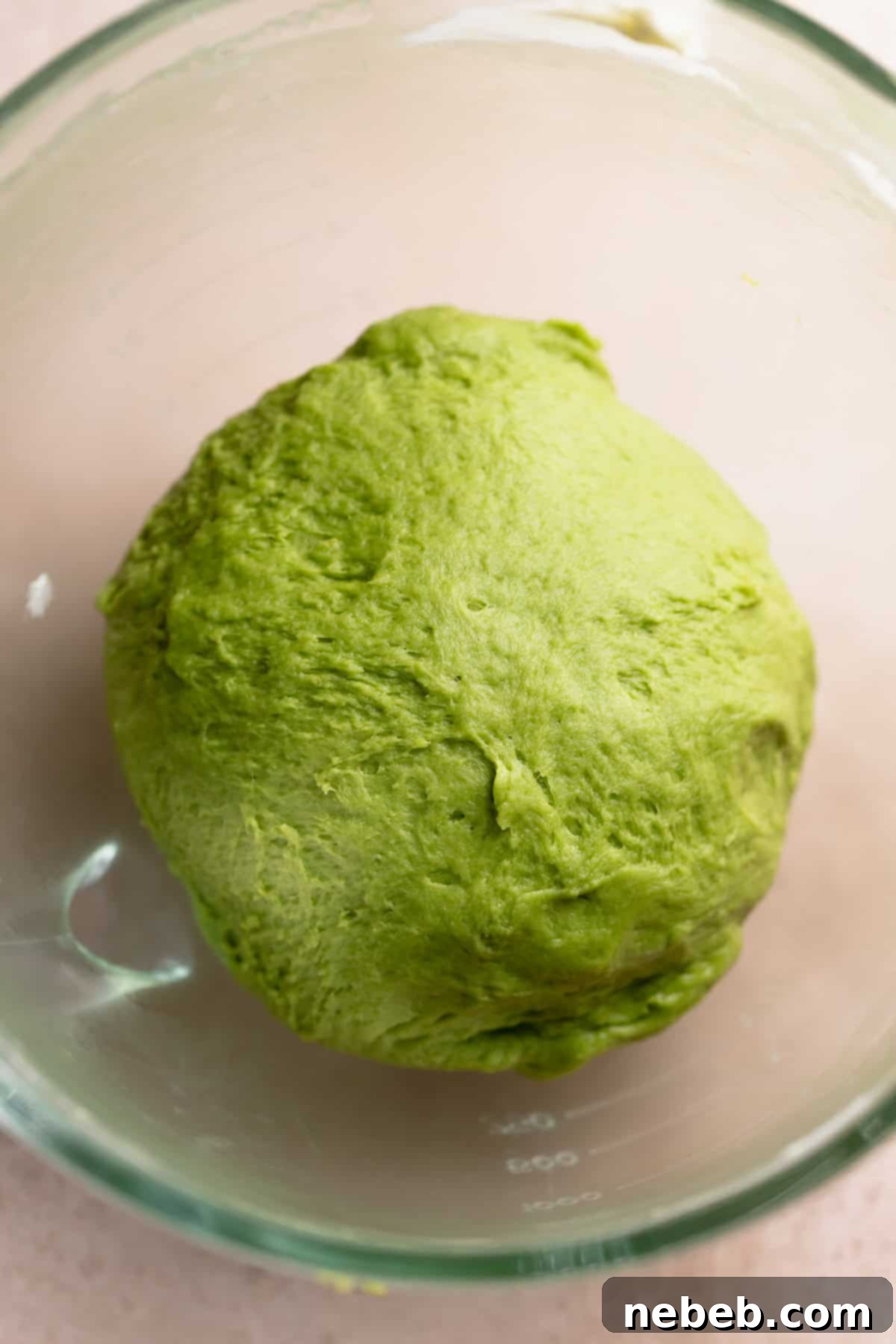 Matcha dough ball resting in a clear glass mixing bowl, covered with plastic wrap.