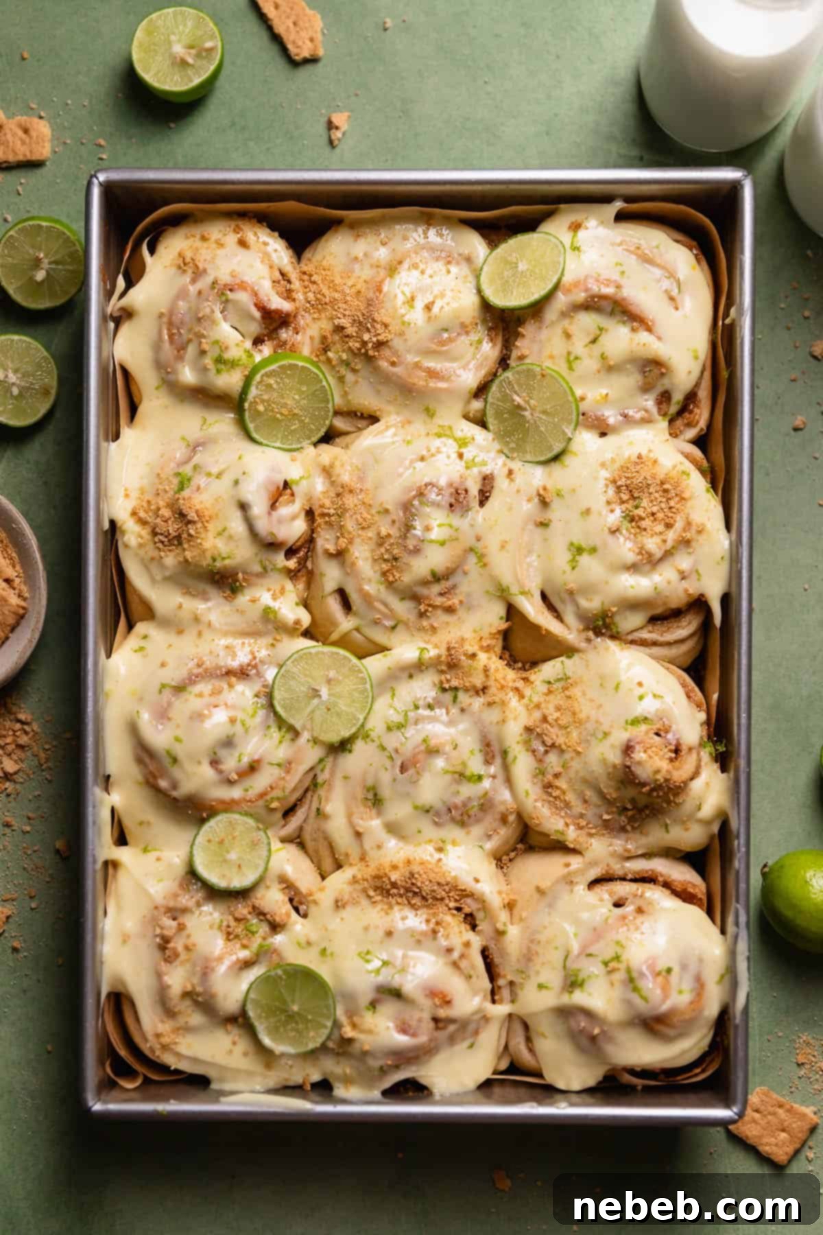 Key lime pie cinnamon rolls in a baking pan with key limes and graham cracker crumbs on top.