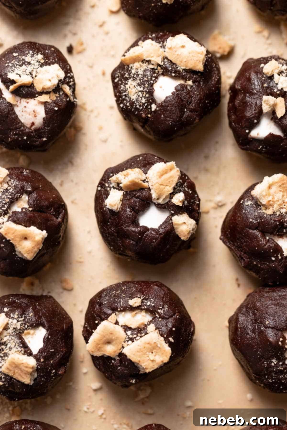 Cookie dough balls, each stuffed with a marshmallow and topped with graham cracker pieces, arranged on a baking tray.