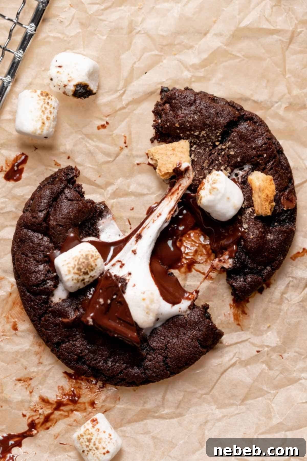 A chocolate s'mores cookie pulled in half on parchment paper, revealing a stretchy, gooey marshmallow center.