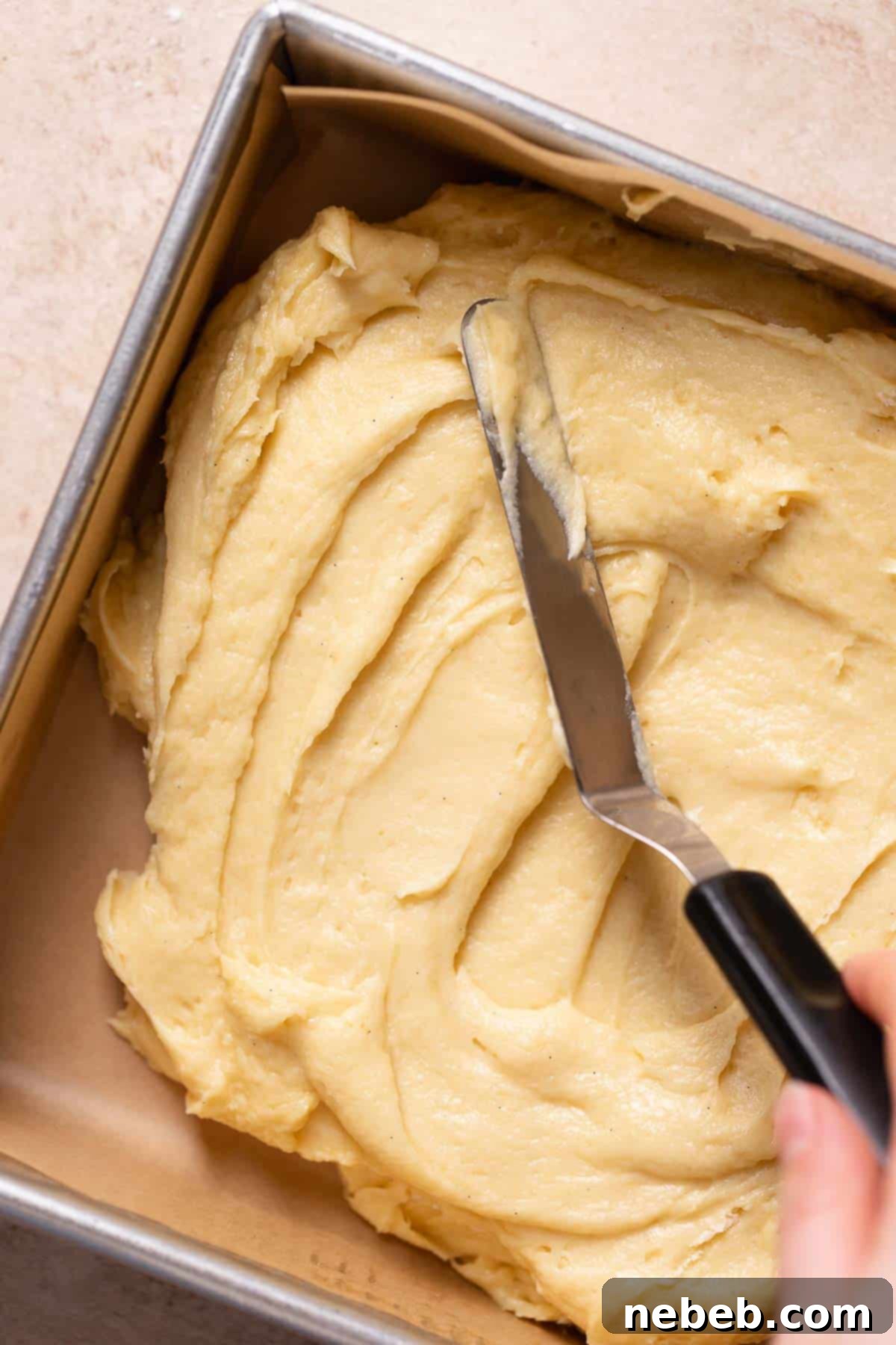 A spatula gently spreading vanilla cake batter into a square baking pan.